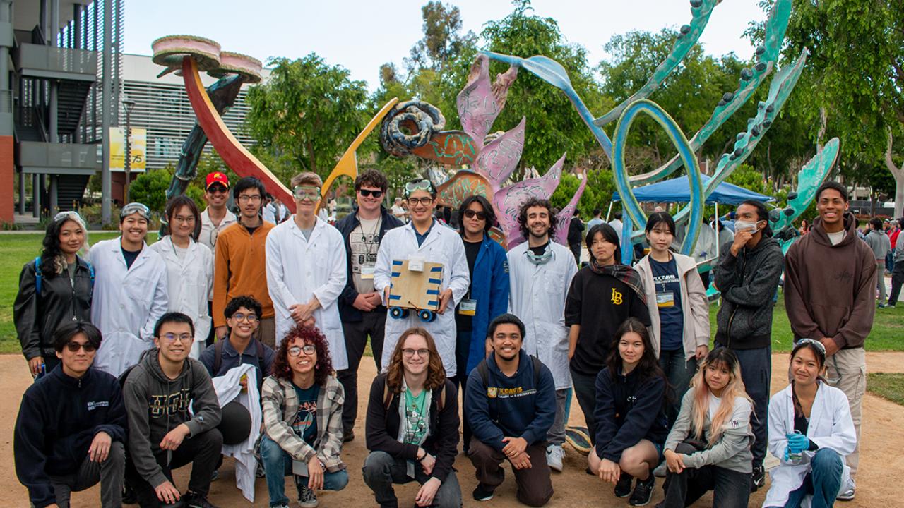 A large group of Chem-E-Car members stand together outdoors near a sculpture, one person in the middle holds the car