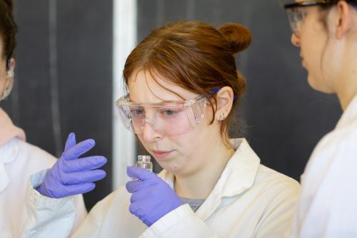 Student wearing safety googles and gloves holding a vial close to their face while wafting the smell closer to her.