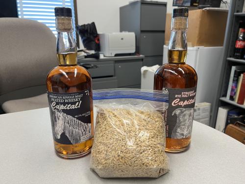 Two bottles of whiskey with labels that say "American Single Malt Peated Whisky Capital!" and a single bag of barley sit on a table.